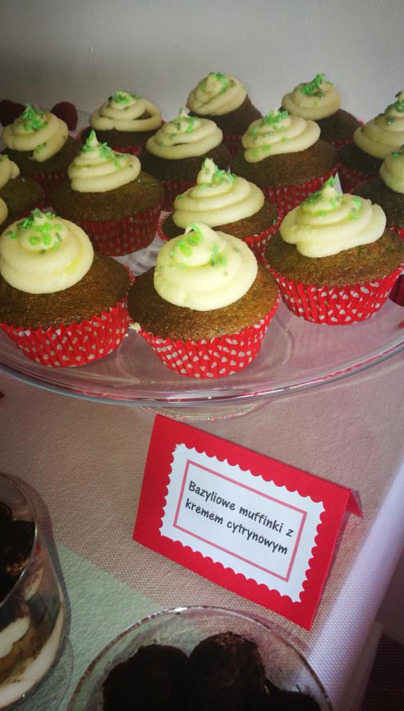 Muffiny bazyliowe z kremem cytrynowym w czerwonych papilotkach na szklanej paterze, udekorowane kremem i zielonymi posypkami, obok tabliczka z nazwą deseru i inne słodkości w szklanych naczyniach.