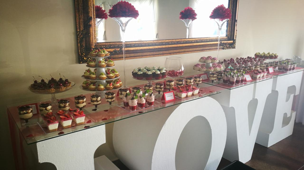 Stół z deserami na przyjęciu we Wrocławiu, ustawiony na białych, dużych literach tworzących napis LOVE, z różnorodnymi słodkościami: tartaletki z owocami, babeczki, desery w szklankach, całość...