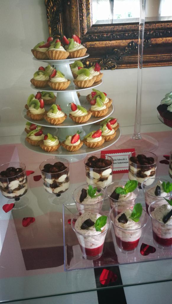 Elegancko zaprezentowane desery: tartaletki z kremem i owocami na trzypiętrowym stojaku oraz desery warstwowe w przezroczystych pucharkach, dekorowane listkami mięty, na błyszczącym stole...