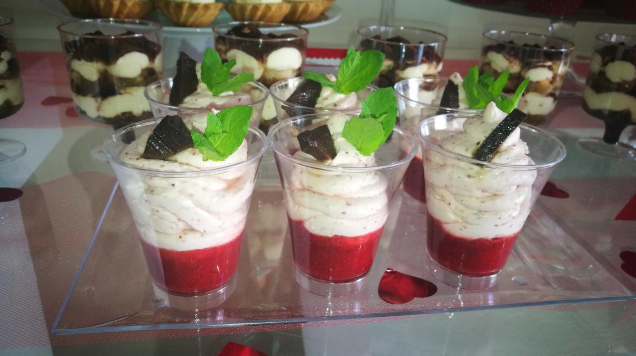 Deser w przezroczystym kubeczku: warstwa czerwonego musu na dole, kremowa pianka na górze, udekorowany listkiem mięty i kawałkiem ciemnej czekolady, na stole z dekoracjami w kształcie serc.