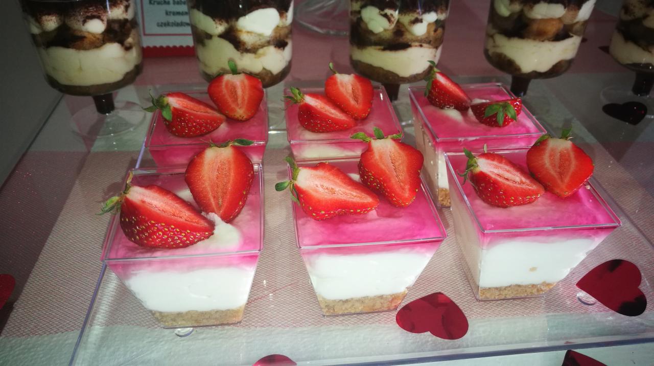 Deser w przezroczystych, kwadratowych pojemniczkach z warstwą ciasteczkowego spodu, białej masy i różowej galaretki, udekorowany połówkami świeżych truskawek; w tle widoczne desery w wysokich...