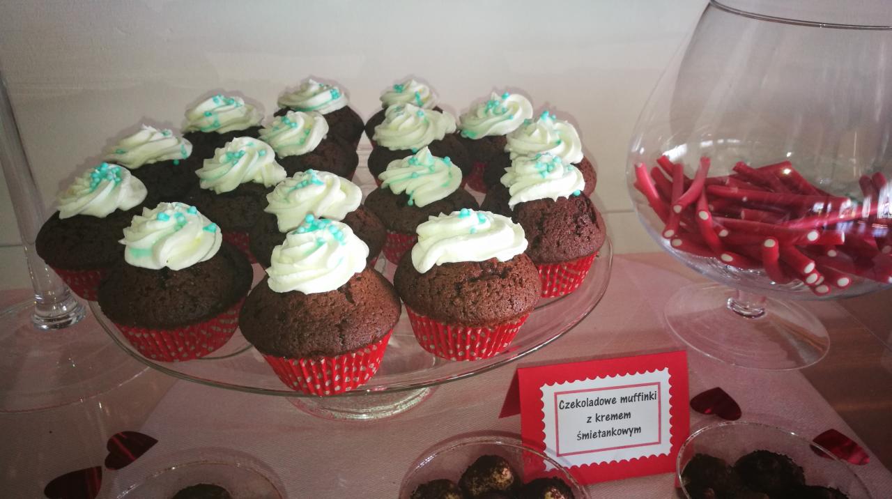 Szklany pater z czekoladowymi muffinami w czerwonych papilotach, udekorowanymi jasnym kremem i turkusowymi posypkami, obok szklane naczynie z czerwonymi żelkami i karteczka z napisem 'Czekoladowe...