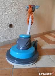 Profesjonalna, niebiesko-szara maszyna do polerowania podłóg z pomarańczowym kablem zwiniętym na uchwycie, stojąca na kafelkach wewnątrz pomieszczenia.