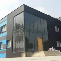 Nowoczesny budynek biurowy z elewacją w kolorach szarości i błękitu, dużą przeszkloną fasadą i surowym drewnianym wejściem, widoczne worki z materiałami budowlanymi na schodach.