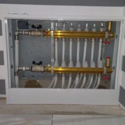 Rozdzielacz ogrzewania podłogowego w metalowej szafce, widoczne rury doprowadzające wodę i liczne węże do pętli grzewczych, na pierwszym planie jasna podłoga z płytek.