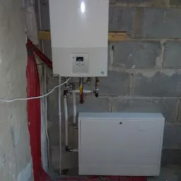 Nowoczesny, biały piec gazowy marki Vaillant zamontowany na surowej ścianie z betonowych pustaków, z widocznymi czerwonymi rurami ogrzewania podłogowego i skrzynką rozdzielczą poniżej.