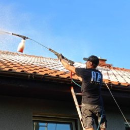 AZ Clean Mycie i malowanie dachów, czyszczenie elewacji, kostki brukowej - Pracownik na drabinie czyści dach z czerwonej dachówki za pomocą myjki ciśnieniowej z pojemnikiem na detergent, widok z dołu na tle błękitnego nieba.
