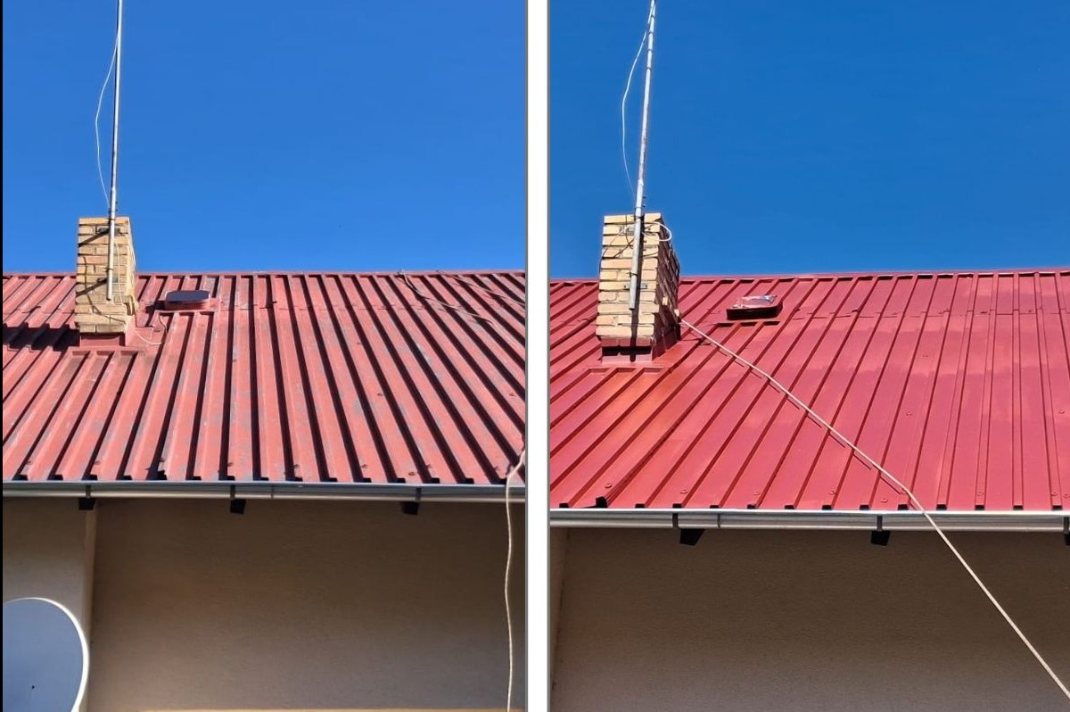 Czerwony dach z blachy trapezowej po renowacji, widoczny komin z cegły i antena. Ujęcie z dołu, błękitne niebo w tle, rynna wzdłuż ściany budynku.