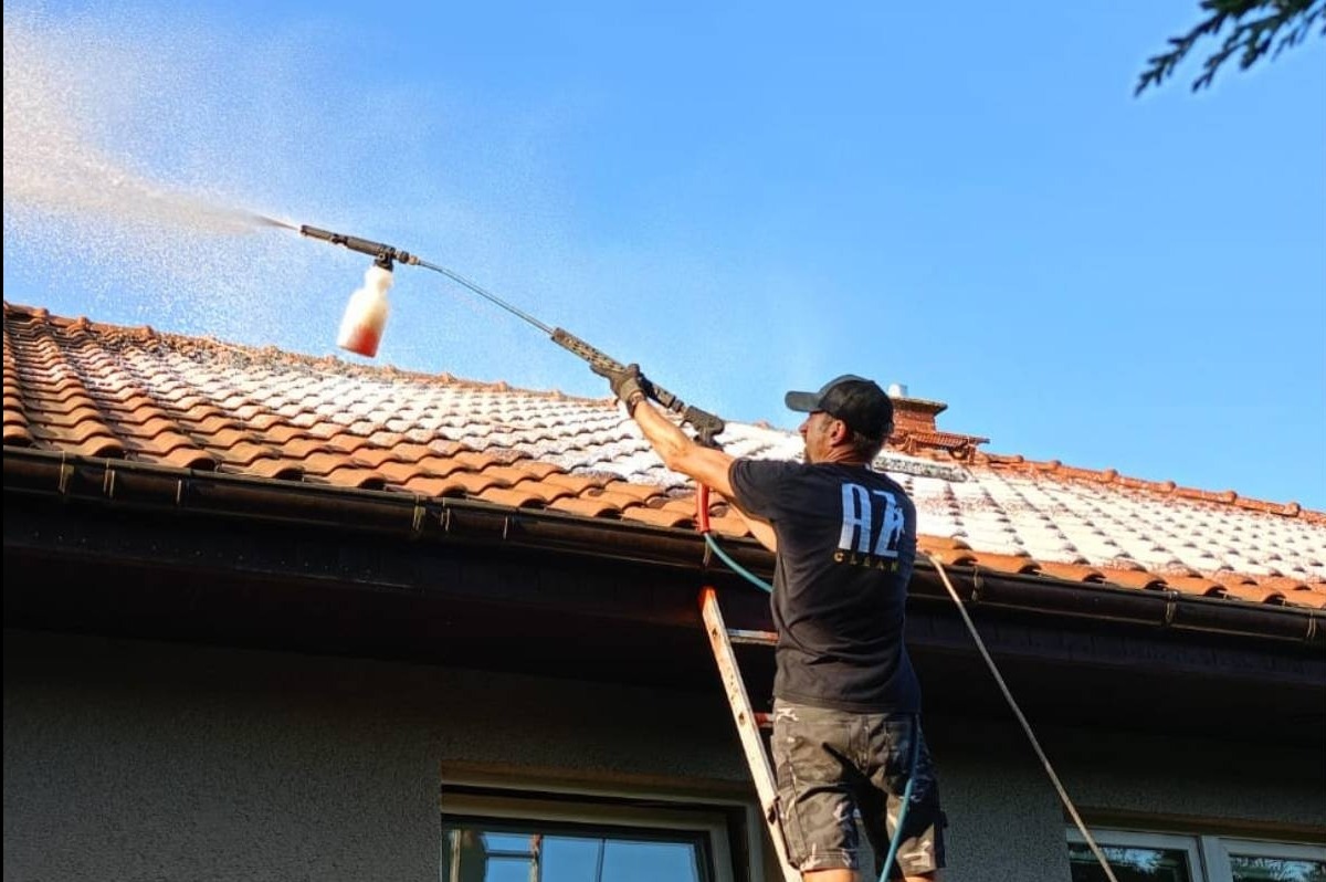 Pracownik na drabinie czyści dach z czerwonej dachówki za pomocą myjki ciśnieniowej z pojemnikiem na detergent, widok z dołu na tle błękitnego nieba.