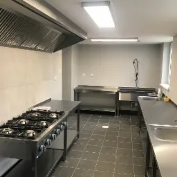 Profesjonalna kuchnia ze stali nierdzewnej z dużą kuchenką gazową, okapem i zlewozmywakami, wykończona szarą glazurą i ciemną podłogą.