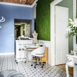 Wnętrze salonu kosmetycznego z zieloną ścianą imitującą trawę, białe lilie w wazonie na białej konsoli, stanowisko do manicure z lampą i krzesłem, manekin krawiecki w tle.