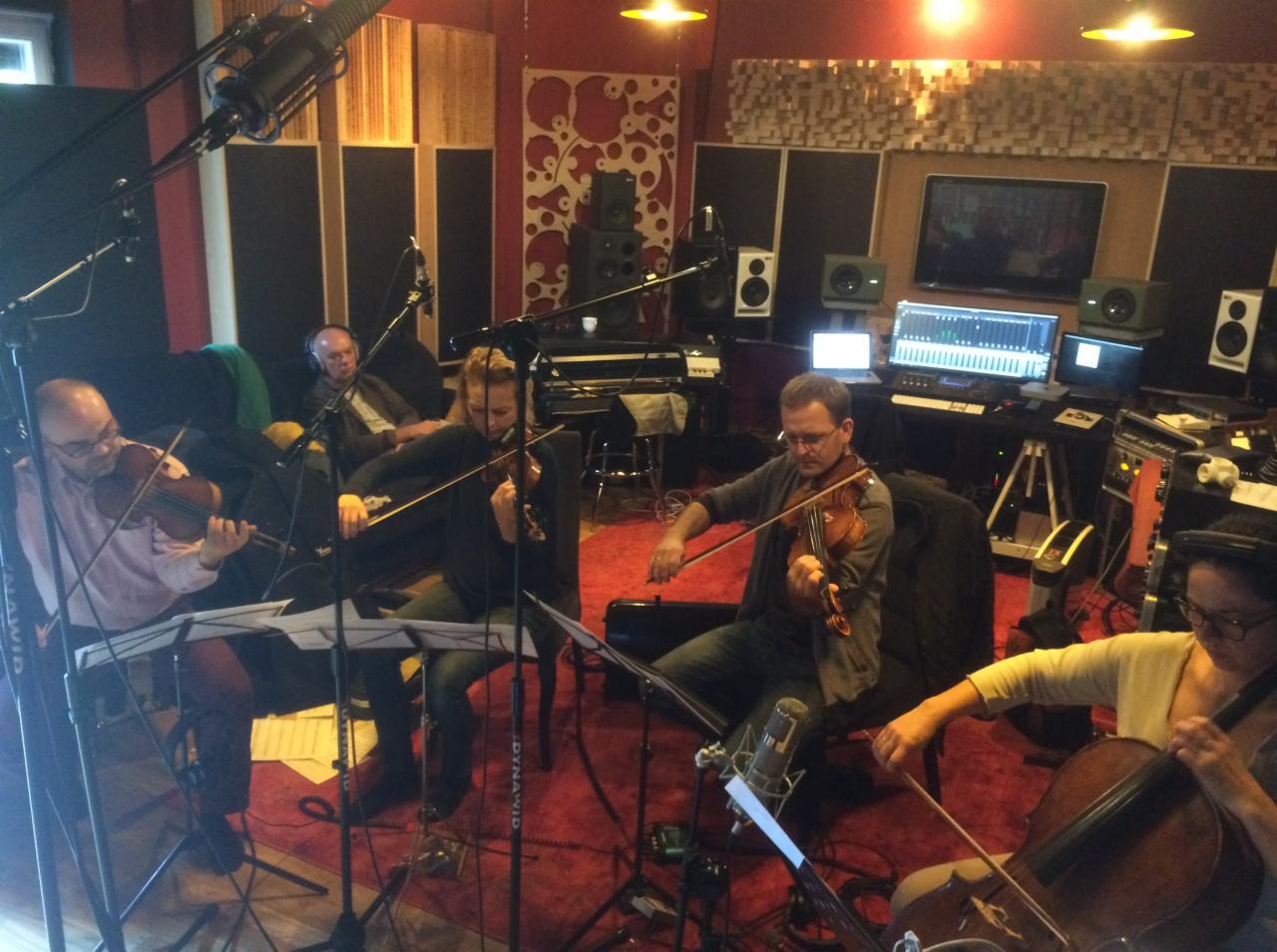 Kwartet smyczkowy nagrywa utwór w profesjonalnym pomieszczeniu z mikrofonami, ekranami, instrumentami klawiszowymi i panelami akustycznymi na ścianach.