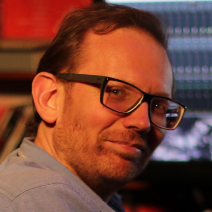 Portret mężczyzny w okularach na tle sprzętu audio, lekki uśmiech, nieostre tło.