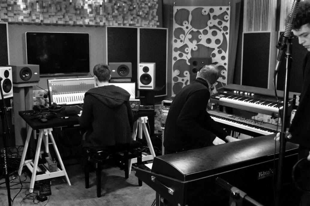 Czarno-białe zdjęcie przedstawia dwóch mężczyzn pracujących przy sprzęcie muzycznym: jeden siedzi przed komputerem z oprogramowaniem DAW, drugi gra na klawiszach. W tle monitory studyjne i panele...