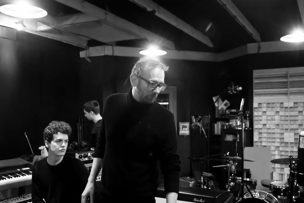 Czarno-białe zdjęcie przedstawia dwóch mężczyzn w pomieszczeniu z instrumentami muzycznymi, w tle widoczne elementy wyposażenia i mikrofony.