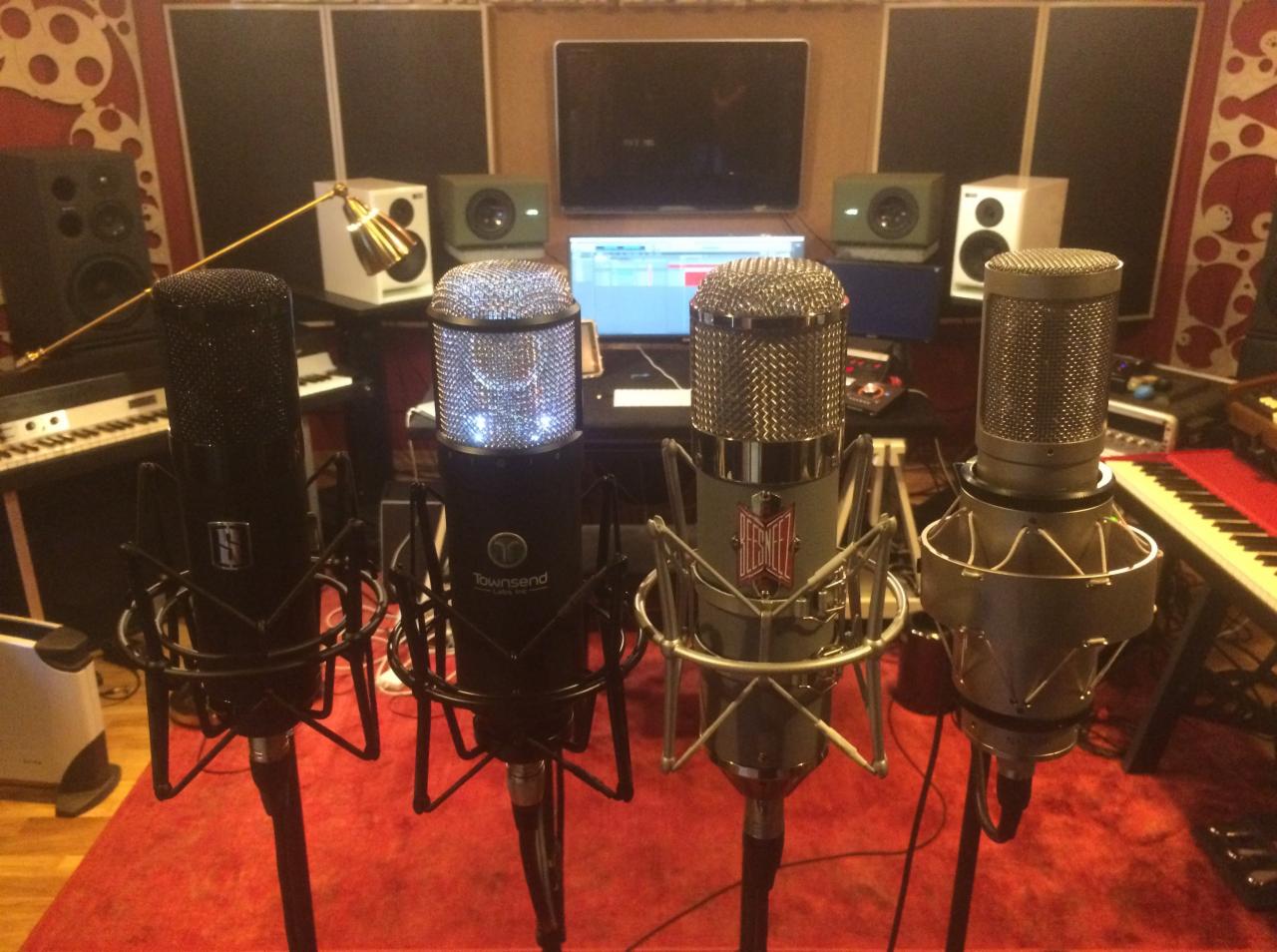 Cztery mikrofony studyjne różnych marek (Townsend, BeesNeez) ustawione w rzędzie na czerwonym dywanie w profesjonalnym studiu nagrań z widocznymi monitorami studyjnymi, klawiaturami i ekranem...