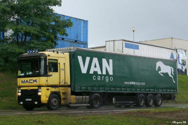 Żółta ciężarówka marki MACK z zieloną naczepą VAN Cargo, na tle magazynów i drzew.