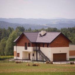 JC Dom - Dwukondygnacyjny dom z drewnianą elewacją na tle zalesionych wzgórz, widoczne schody zewnętrzne i baloty siana na polu.