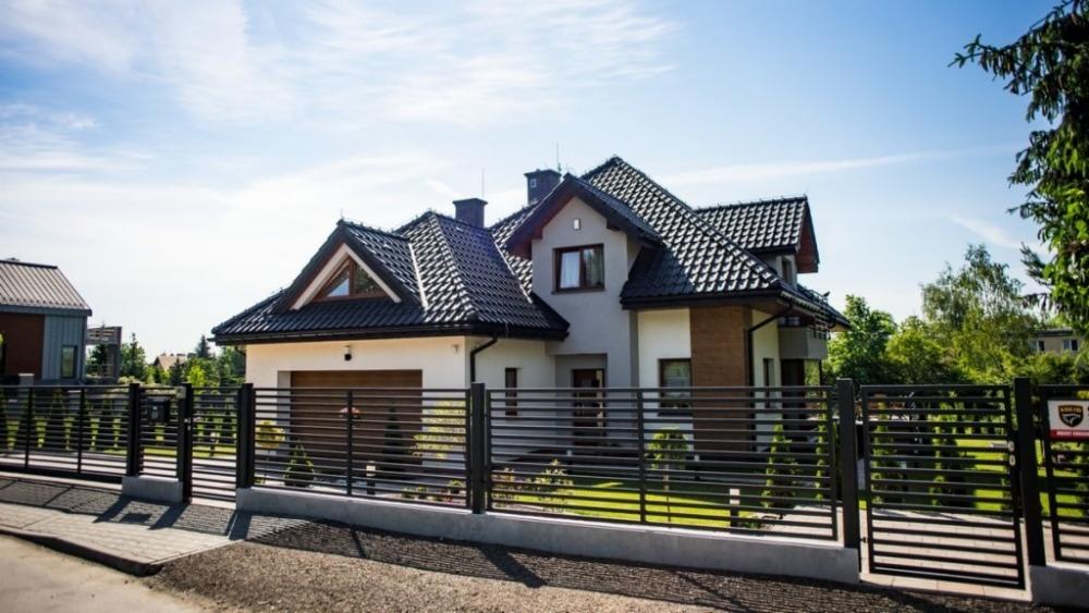 Nowoczesny dom jednorodzinny z ciemną dachówką i jasną elewacją, ogrodzony metalowym płotem, widoczny z poziomu ulicy w słoneczny dzień.