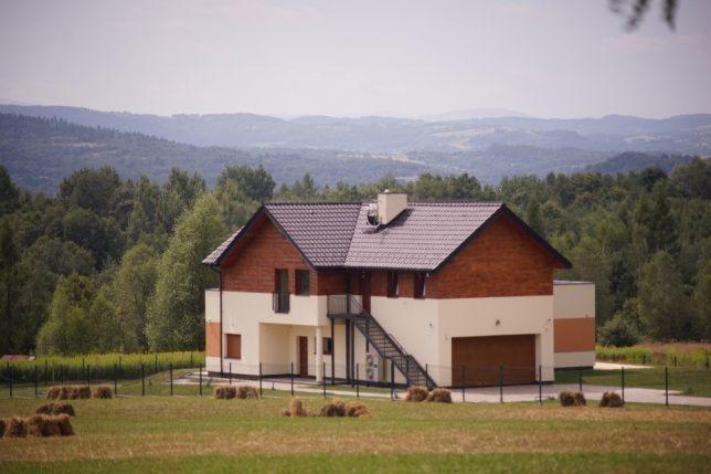 Dwukondygnacyjny dom z drewnianą elewacją na tle zalesionych wzgórz, widoczne schody zewnętrzne i baloty siana na polu.