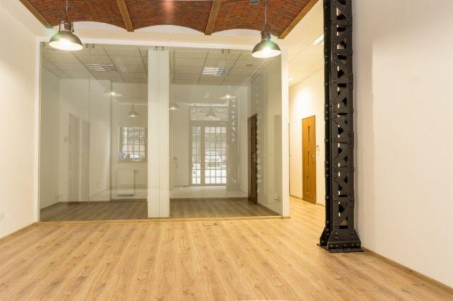 Przestronne, puste wnętrze w stylu loft z drewnianą podłogą, szklaną ścianą działową i odsłoniętą, dekoracyjną stalową kolumną.