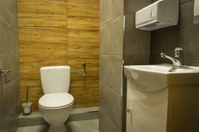 Nowoczesna łazienka z drewnopodobnymi płytkami na ścianie, białą toaletą i umywalką z szafką, oraz podajnikiem na ręczniki papierowe.