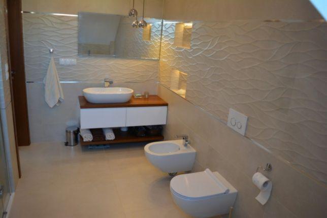 Nowoczesna łazienka z umywalką nablatową na drewnianym blacie, bidetem i toaletą w jasnych odcieniach, wyróżniająca się falistą strukturą płytek ściennych i dekoracyjnym oświetleniem.