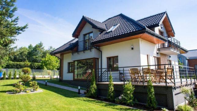 Dwukondygnacyjny dom z białymi ścianami, ciemnym dachem, balkonem z drewnianymi krzesłami i zadbanym trawnikiem z ozdobnymi krzewami.