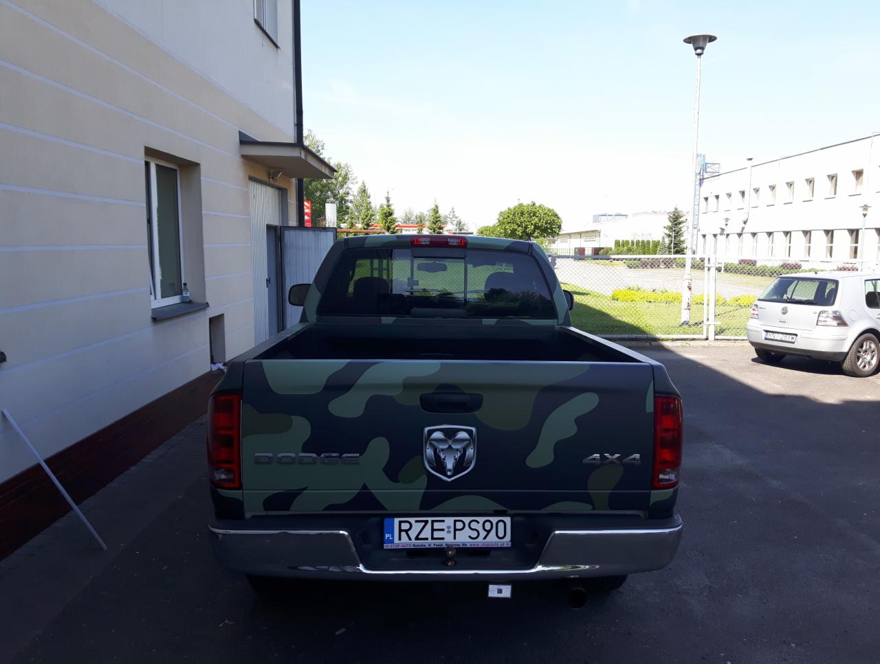 Pojazd Dodge Ram z kamuflażem w odcieniach zieleni, widok z tyłu, na parkingu przed budynkiem.