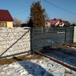 Przesuwna brama wjazdowa z poziomych profili, zintegrowana z gabionowym ogrodzeniem wypełnionym białym kamieniem, widok na posesję z domami w tle, częściowo pokrytą śniegiem.