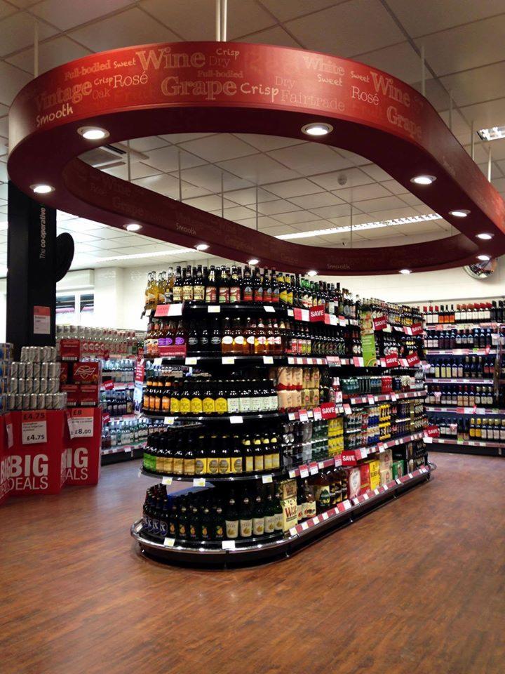 Zdjęcie przedstawia zakrzywiony regał sklepowy z różnymi butelkami wina, nad którym znajduje się dekoracyjny element sufitowy z napisem 'Wine Grape Rose', wykonany z drewna w odcieniach czerwieni...