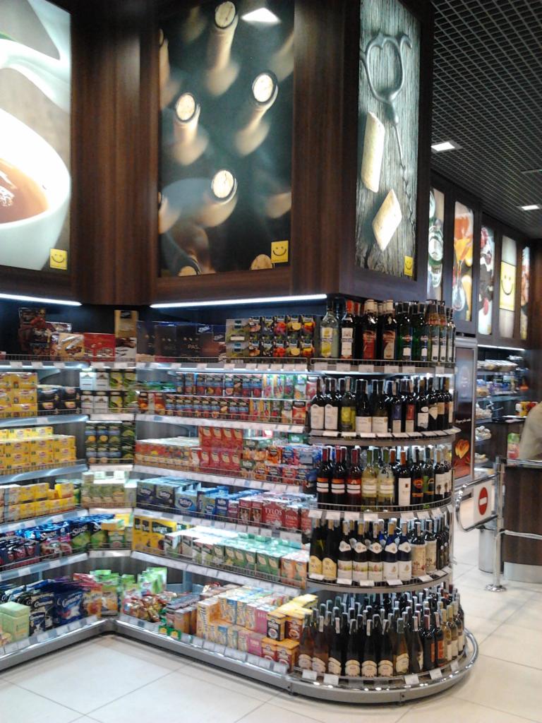 Wnętrze sklepu spożywczego z półkami wypełnionymi różnorodnymi produktami, w tym butelkami wina na obrotowej półce, z dekoracyjnymi panelami na górze przedstawiającymi wina i jedzenie.