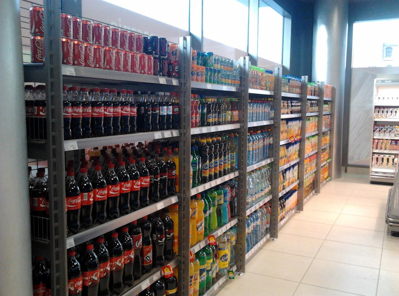 Metalowe regały sklepowe wypełnione napojami gazowanymi i wodami mineralnymi, widok z perspektywy klienta w jasnym wnętrzu sklepu.