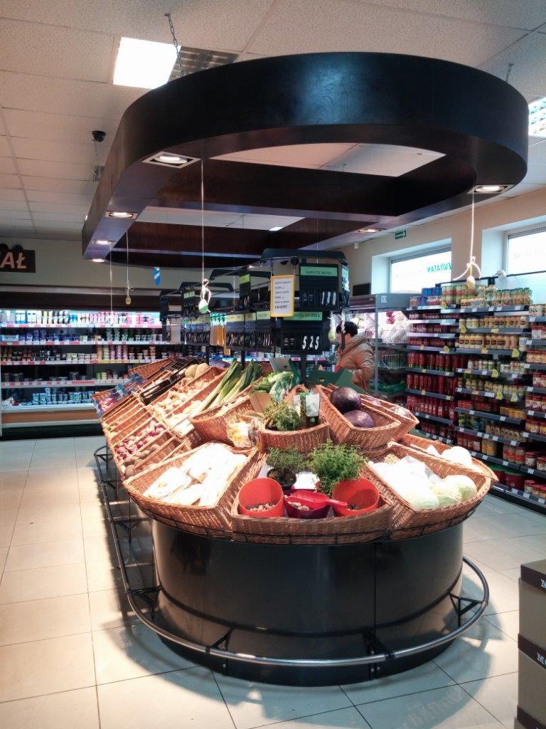 Ekspozycja warzyw i owoców w koszach wiklinowych na okrągłym stoisku w supermarkecie, pod ciemnym, drewnianym baldachimem z oświetleniem punktowym.
