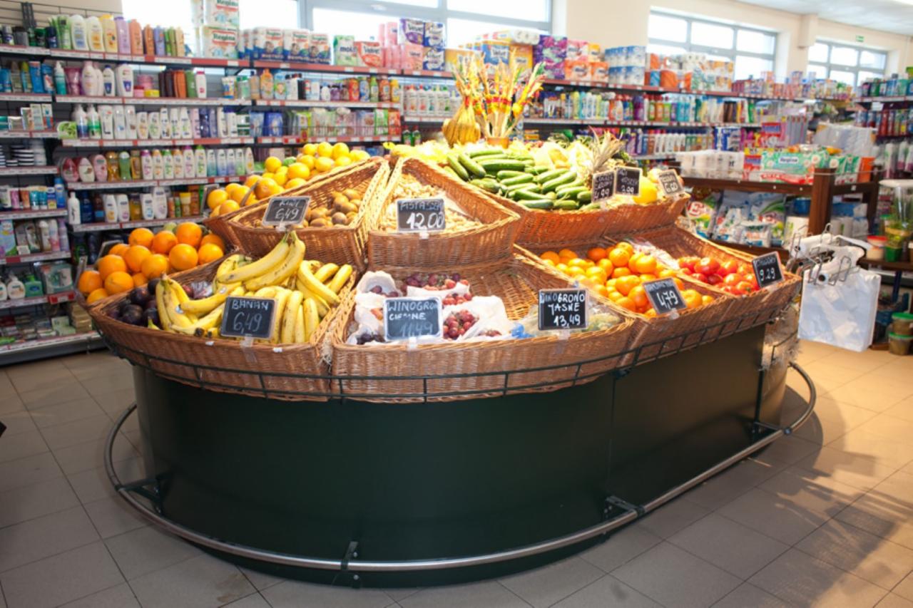 Ekspozycja owoców i warzyw w sklepie spożywczym: banany, pomarańcze, kiwi, ogórki i winogrona w wiklinowych koszach z cenami na tle regałów z towarem.