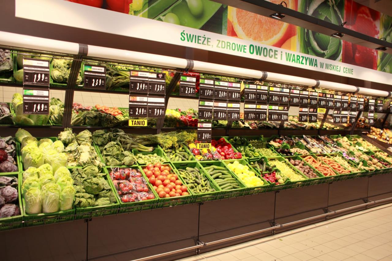 Rząd zielonych skrzynek wypełnionych świeżymi warzywami w supermarkecie, z cenami zawieszonymi nad każdym produktem i hasłem reklamowym na górze.
