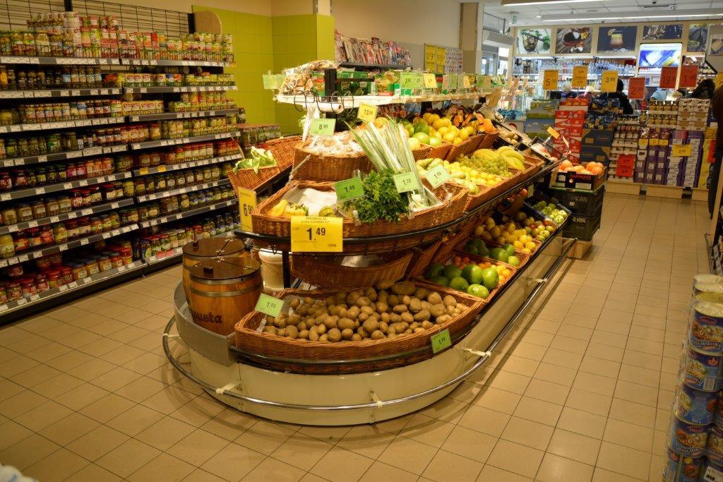 Ekspozycja warzyw i owoców w sklepie spożywczym, z ziemniakami, zieleniną i owocami ułożonymi w koszach na wielopoziomowym stojaku, obok regały z konserwami i innymi produktami.