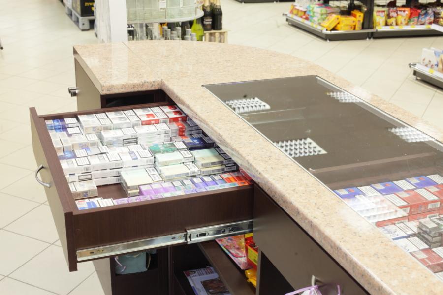 Brązowa lada sklepowa z marmurowym blatem i otwartą szufladą wypełnioną różnymi paczkami papierosów; widoczne metalowe szyny szuflady i fragment półki pod szufladą z kolorowymi opakowaniami.