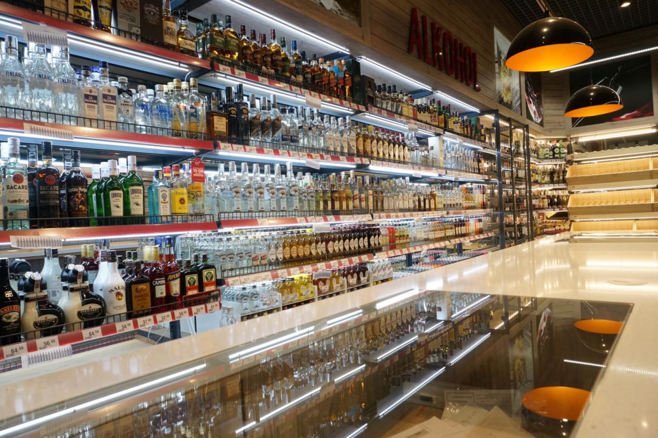 Półki sklepowe wypełnione różnorodnymi butelkami alkoholi, z podświetleniem LED i odblaskiem w blacie lady, tworzące wrażenie głębi i obfitości.