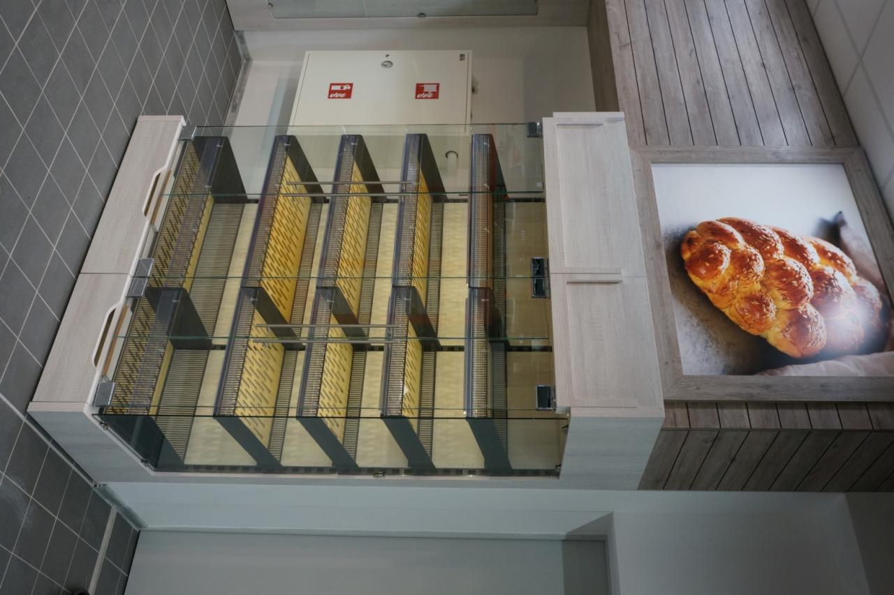 Wnętrze piekarni z przeszkloną ekspozycją pieczywa, widoczna grafika chleba na ścianie, szara podłoga i drewnopodobna okładzina ściany.