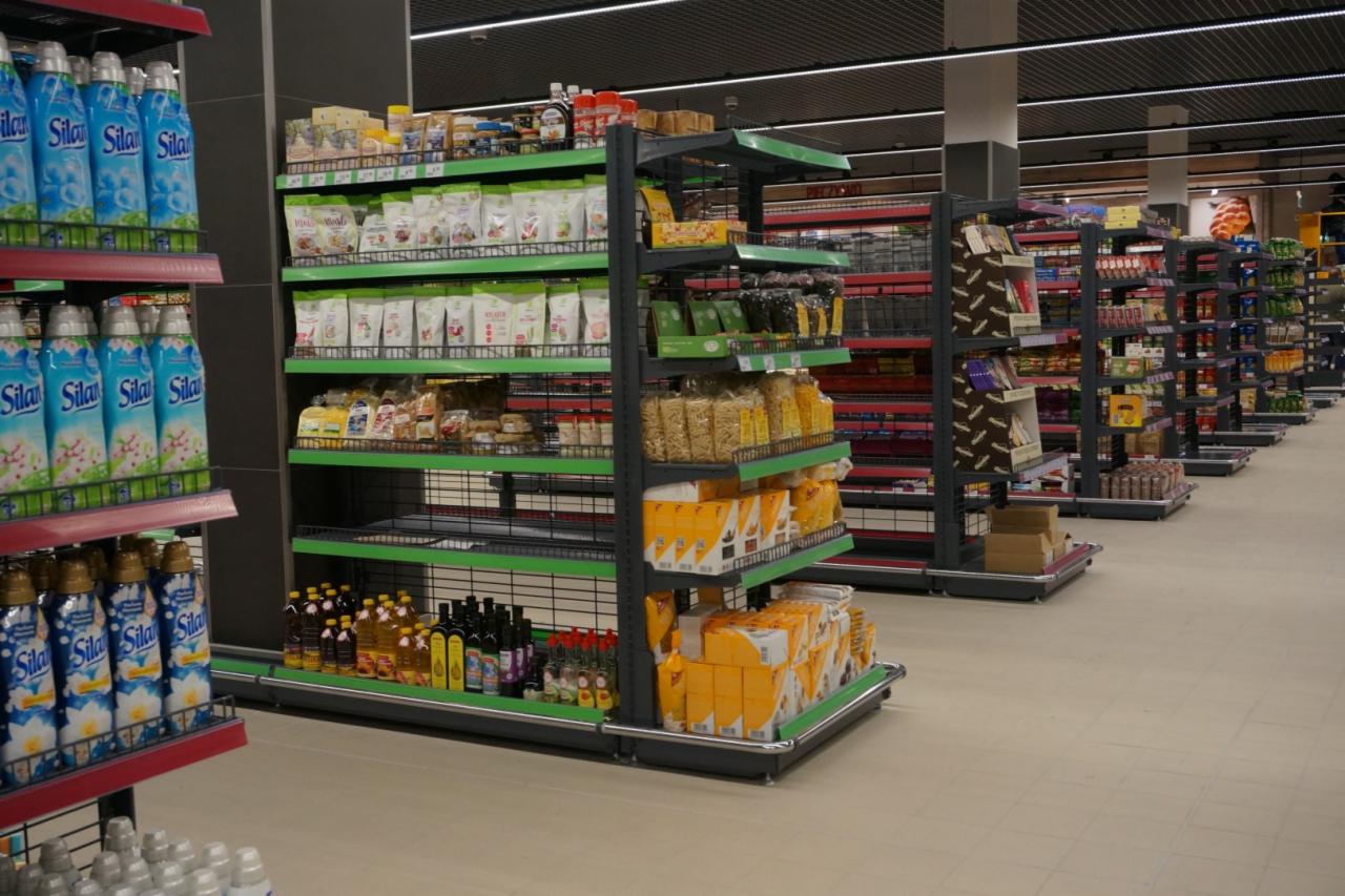 Półki sklepowe z metalowymi regałami w odcieniach szarości i zieleni, wypełnione różnorodnymi produktami spożywczymi i chemii gospodarczej, widok w perspektywie w głąb sklepu.