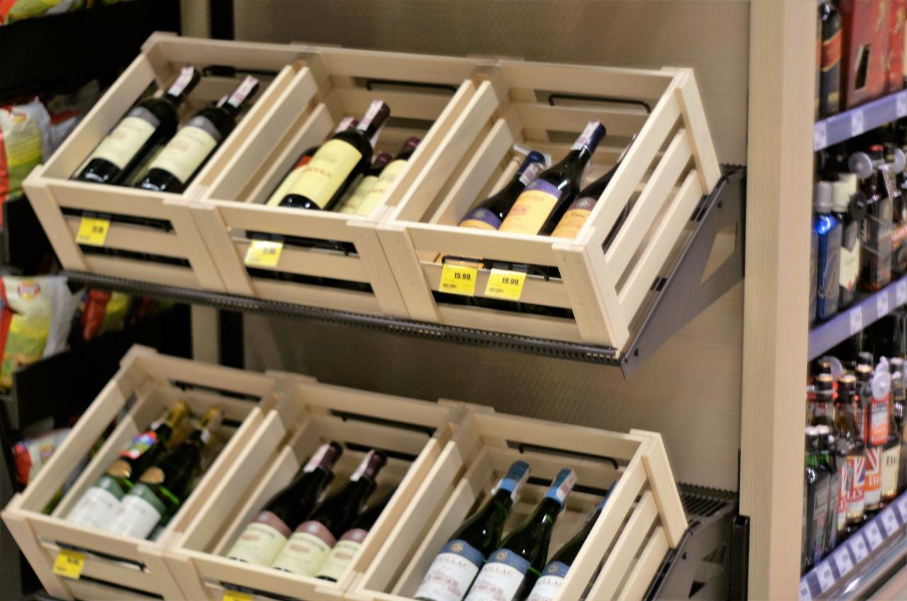 Drewniane skrzynki na wino zamontowane na metalowych prowadnicach w sklepie, z cenami na każdej skrzynce, widok z boku na regał z innymi alkoholami.