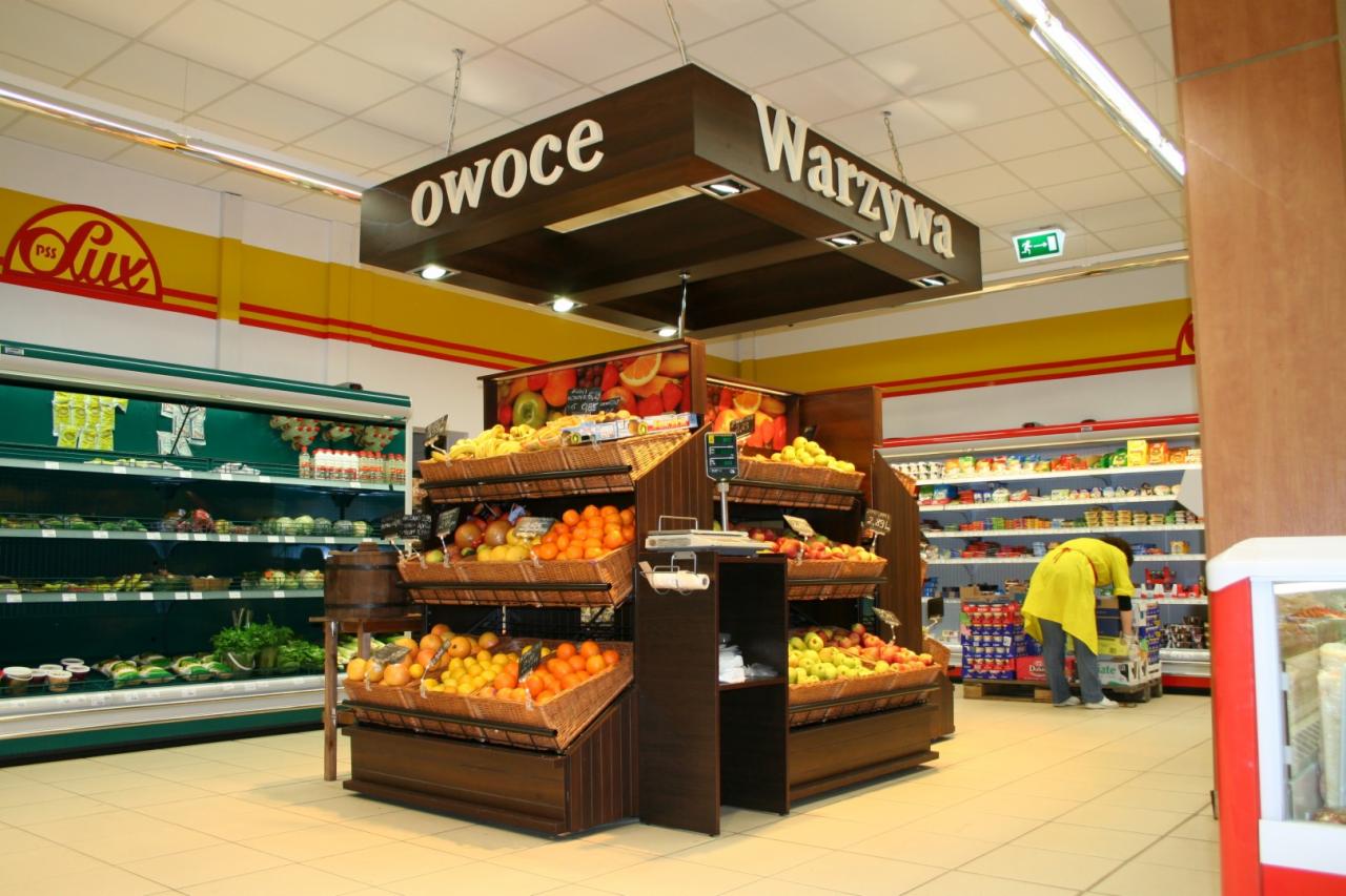 Drewniany regał ekspozycyjny na owoce i warzywa w supermarkecie, z podświetlanym napisem 'Owoce' i 'Warzywa' nad nim, kosze wiklinowe z pomarańczami, jabłkami i innymi owocami, w tle chłodnia...