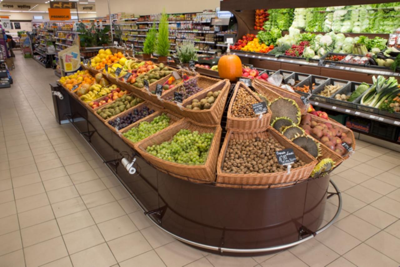Ekspozycja owoców i warzyw w supermarkecie, z koszami wiklinowymi zawierającymi winogrona, kiwi, orzechy laskowe, pomarańcze, banany i inne, z dynią na górze, na tle regałów sklepowych.