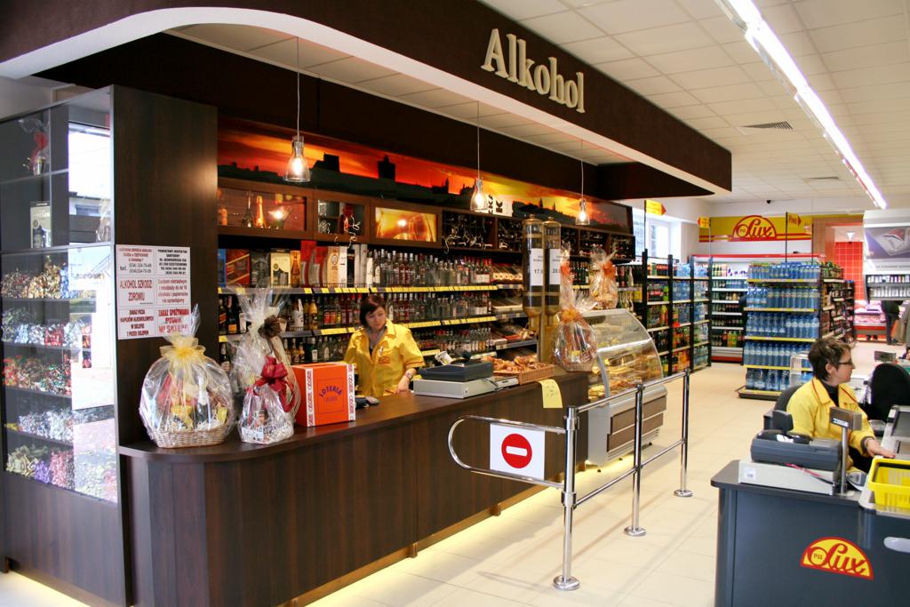 Widok wnętrza sklepu z alkoholem, z półkami pełnymi butelek, kasą obsługiwaną przez kasjerkę w żółtej bluzce, dekoracyjnymi koszami prezentowymi i napisem 'Alkohol' nad stoiskiem.