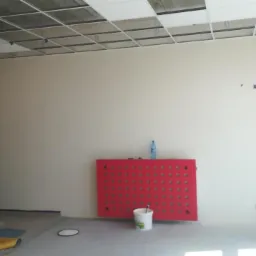 Świeżo pomalowane wnętrze biurowe z widocznym sufitem podwieszanym, czerwonymi akcentami okiennymi i rolkami folii malarskiej w rogu. Na podłodze widoczne ślady farby i wiadro z narzędziami.