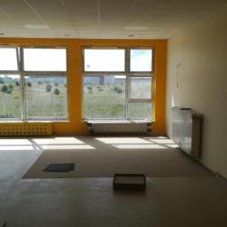 Reaktart - Wnętrze pokoju z żółtymi ramami okiennymi i grzejnikami, przygotowane do prac wykończeniowych na podłodze, widoczne fragmenty podkładu i kartonowe pudełko.