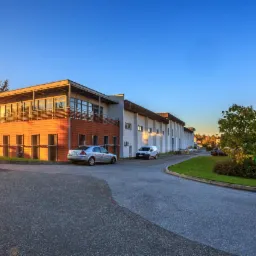 Nowoczesny budynek usługowy z pomarańczową elewacją i balkonem, oświetlony zachodzącym słońcem, z zaparkowanymi samochodami na parkingu.