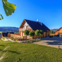 Nowoczesny, drewniany dom z balkonem, czarnym dachem i satelitą, otoczony zielonym trawnikiem i drzewami, z widokiem na góry w tle, sfotografowany w słoneczny dzień.