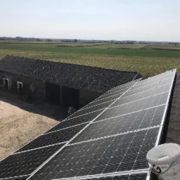 Panel słoneczny na dachu domu z widokiem na wiejskie pole, z wiaderkiem farby obok paneli, w tle budynek gospodarczy.